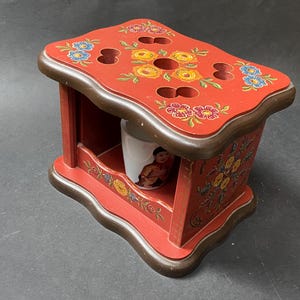 May include: A red wooden box with a decorative floral design and a small white cup inside. The box has a scalloped edge and is painted with flowers in shades of red, yellow, blue, and green.