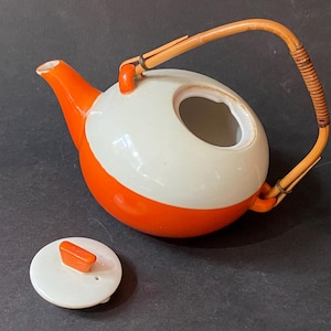 Könnte beinhalten: Eine weiße und orangefarbene Teekanne aus Keramik mit einem Bambusgriff und einem separaten Deckel. Die Teekanne hat einen runden Körper und einen Ausguss an der Seite.