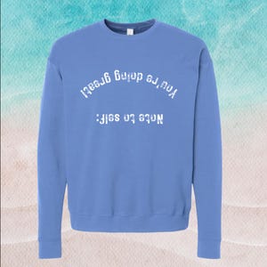 May include: A periwinkle blue crewneck sweatshirt featuring white text printed in an upside-down orientation. The text reads Note to self: You're doing great! The garment has long sleeves, a ribbed neckline, and ribbed cuffs at the wrists and hem.