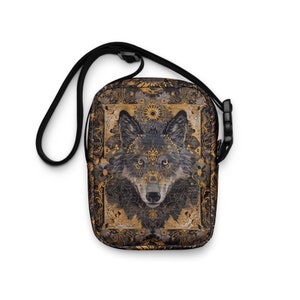 Könnte beinhalten: Schwarze Umhängetasche mit detaillierter Wolfsillustration und goldenen Akzenten. Die Tasche hat einen schwarzen, verstellbaren Riemen und eine Schnalle. Das Wolfsdesign befindet sich auf einem dekorativen, floralen Hintergrund.