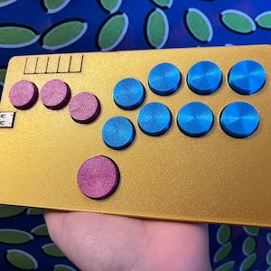 Könnte beinhalten: Ein goldfarbener Arcade-Controller mit einem USB-Anschluss. Der Controller hat acht blaue Knöpfe und vier rosa Knöpfe.