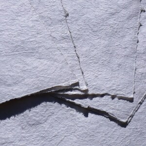 May include: Close-up of handmade, textured paper sheets in shades of white and gray. The paper has rough, deckled edges and a slightly wrinkled surface, suggesting a natural, artisanal quality. Ideal for art projects or stationery.