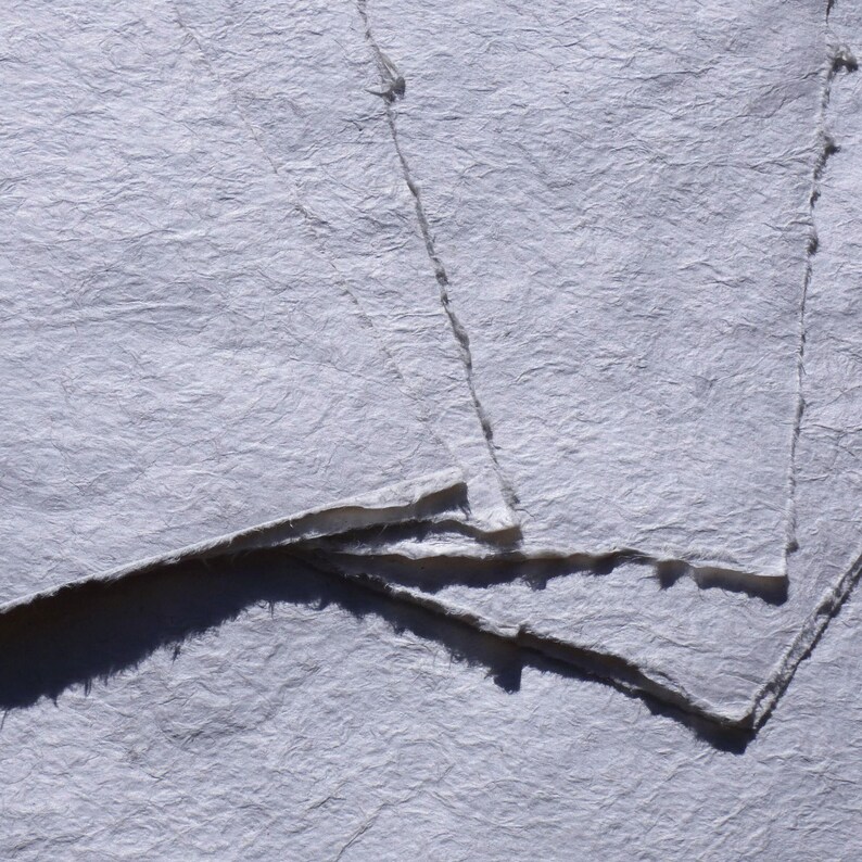 May include: Close-up of handmade, textured paper sheets in shades of white and gray. The paper has rough, deckled edges and a slightly wrinkled surface, suggesting a natural, artisanal quality. Ideal for art projects or stationery.