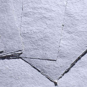 May include: Close-up of several sheets of handmade, textured, light-gray paper. The paper has a rough, uneven surface with visible fibers and a slightly translucent quality. The edges are torn and irregular, suggesting a natural, artisanal process.
