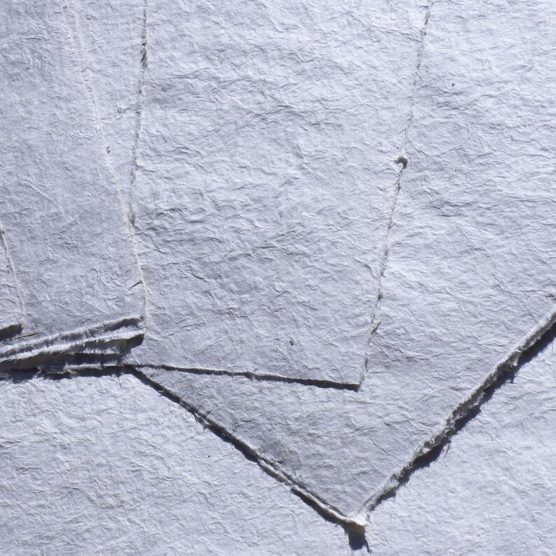 May include: Close-up of several sheets of handmade, textured, light-gray paper. The paper has a rough, uneven surface with visible fibers and a slightly translucent quality. The edges are torn and irregular, suggesting a natural, artisanal process.