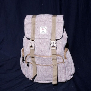 Könnte beinhalten: Ein helllila-beiger Rucksack mit Fischgrätmuster. Der Rucksack hat beige Riemen, Schnallen und Reißverschlüsse. Die obere Klappe hat ein kleines Etikett mit Text. Der Rucksack ist aus gewebtem Material.