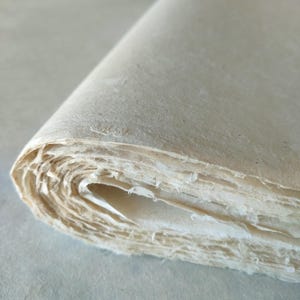 Op de afbeelding: Een opgerolde stapel handgemaakt papier, met de geschepte randen zichtbaar. Het papier heeft een natuurlijke, gebroken witte kleur en een licht getextureerd oppervlak. De lagen zijn zichtbaar, wat een gevoel van diepte en textuur cre&euml;ert. Het papier lijkt dik en van hoge kwaliteit.