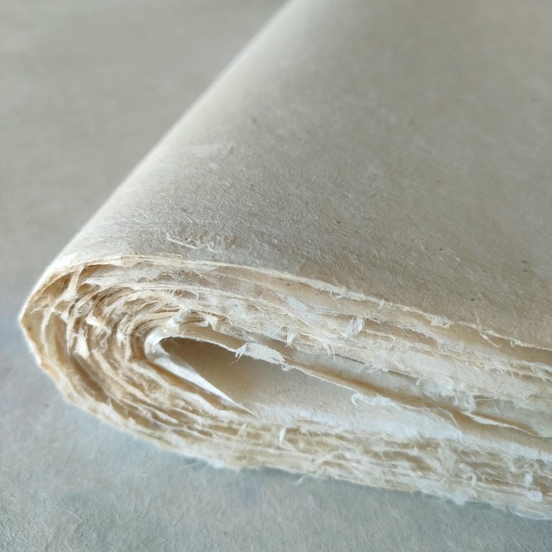 Op de afbeelding: Een opgerolde stapel handgemaakt papier, met de geschepte randen zichtbaar. Het papier heeft een natuurlijke, gebroken witte kleur en een licht getextureerd oppervlak. De lagen zijn zichtbaar, wat een gevoel van diepte en textuur cre&euml;ert. Het papier lijkt dik en van hoge kwaliteit.