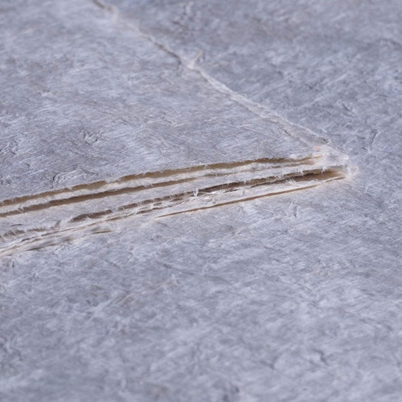 May include: A stack of handmade paper sheets with a textured, fibrous surface. The paper has a light gray color with visible fibers and a slightly rough texture. The edges of the paper are uneven, suggesting a natural, artisanal quality.