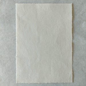 Puede incluir: Una hoja rectangular de papel blanquecino con una superficie texturizada y bordes irregulares. El papel est&aacute; sobre un fondo gris claro. El papel parece hecho a mano, adecuado para proyectos de arte o manualidades.