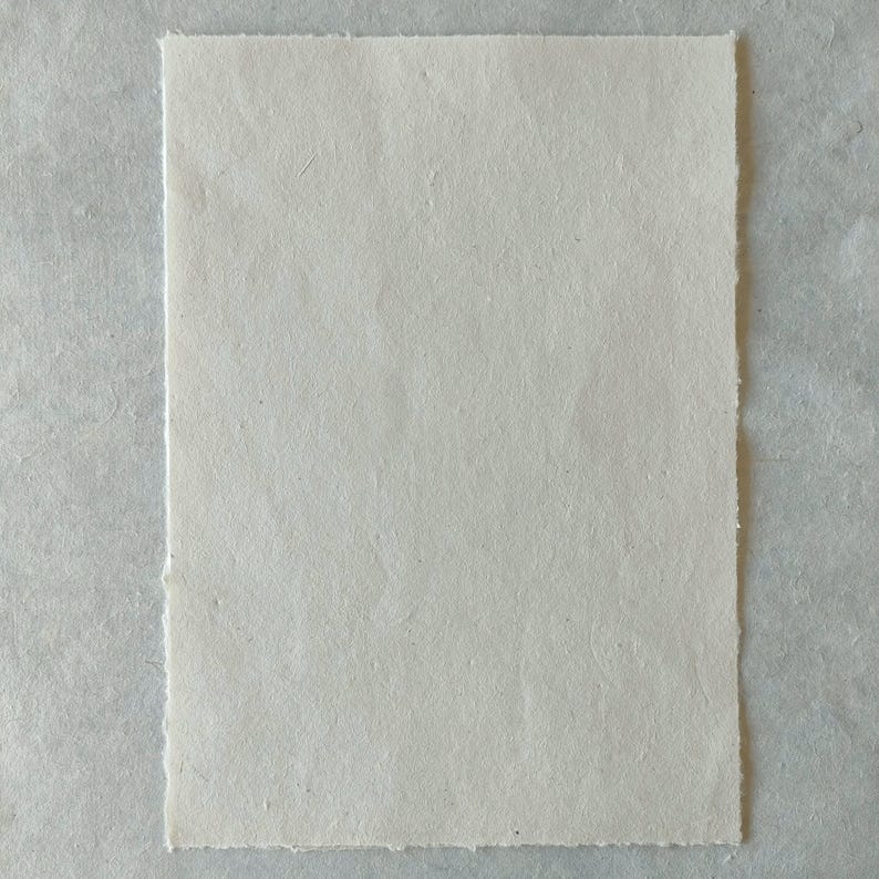 Puede incluir: Una hoja rectangular de papel blanquecino con una superficie texturizada y bordes irregulares. El papel est&aacute; sobre un fondo gris claro. El papel parece hecho a mano, adecuado para proyectos de arte o manualidades.