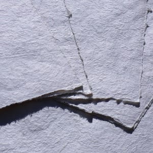 Puede incluir: Primer plano de varias hojas de papel hecho a mano. El papel tiene una superficie texturizada, ligeramente arrugada y un color blanco roto claro. Los bordes del papel son &aacute;speros e irregulares, lo que sugiere una calidad natural y artesanal. El papel est&aacute; apilado.