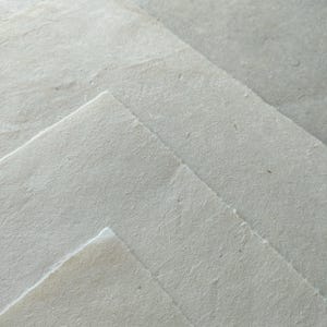 Op de afbeelding: Close-up van verschillende vellen handgemaakt papier. Het papier heeft een ruw, getextureerd oppervlak en een natuurlijke, gebroken witte kleur. De randen van het papier zijn enigszins ongelijk, wat wijst op een ambachtelijk proces. De vellen zijn gelaagd.