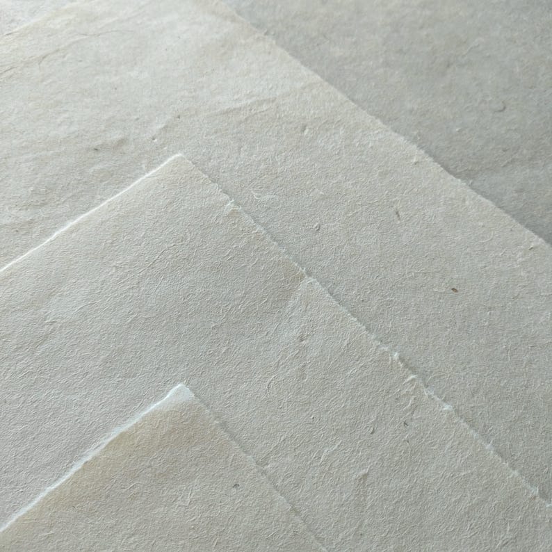 Puede incluir: Primer plano de varias hojas de papel hecho a mano. El papel tiene una superficie rugosa y texturizada y un color blanco roto claro. Los bordes del papel son ligeramente irregulares, lo que sugiere una calidad natural y artesanal. Las hojas est&aacute;n superpuestas, creando profundidad y dimensi&oacute;n.