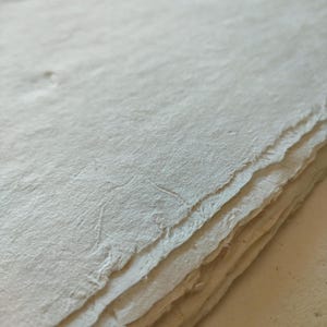 Op de afbeelding: Een stapel handgemaakte, gebroken witte papieren vellen met ruwe, gescheurde randen. Het papier heeft een getextureerd oppervlak en lijkt gemaakt van natuurlijke vezels. De afbeelding is een close-up, die de unieke textuur van het papier benadrukt.