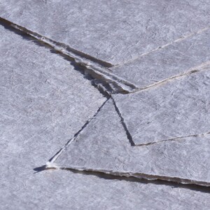 May include: Close-up of several sheets of handmade paper with a textured, fibrous surface. The paper is a pale gray color with deckled edges, creating a rustic and artisanal aesthetic. The sheets are layered, showing the paper's unique texture.