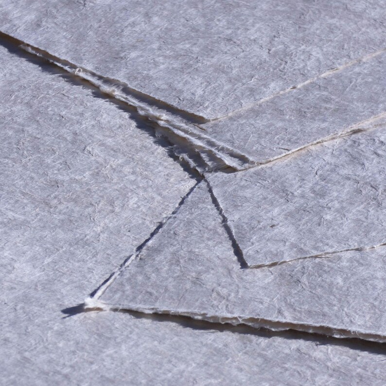 May include: Close-up of several sheets of handmade paper with a textured, fibrous surface. The paper is a pale gray color with deckled edges, creating a rustic and artisanal aesthetic. The sheets are layered, showing the paper's unique texture.
