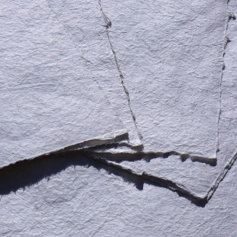 Pu&ograve; includere: Primo piano di diversi fogli di carta fatta a mano. La carta ha una superficie strutturata, leggermente rugosa e un colore bianco sporco chiaro. I bordi della carta sono ruvidi e irregolari, suggerendo una qualit&agrave; naturale e artigianale. La carta &egrave; impilata.