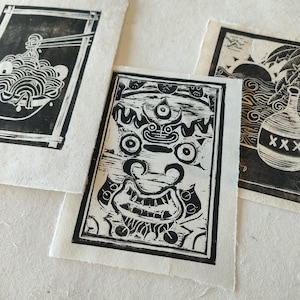 Trois linogravures en noir et blanc sur papier Lokta. La première estampe représente un bol de nouilles avec des baguettes. La deuxième estampe montre un visage stylisé détaillé avec un troisième œil. La troisième estampe montre une bouteille avec le mot "XXX" dessus.