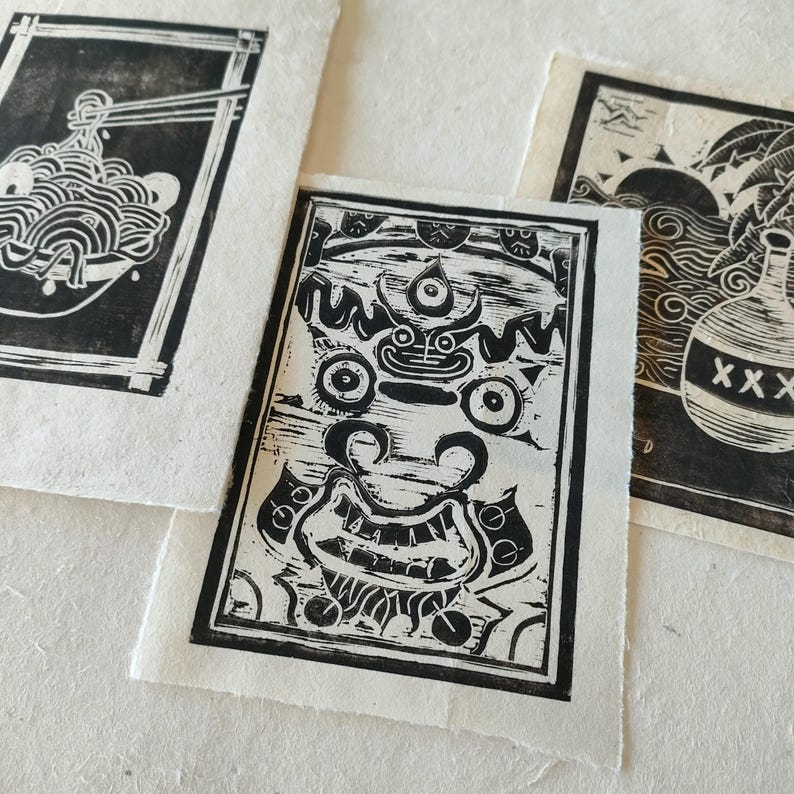 Trois linogravures en noir et blanc sur papier Lokta. La première estampe représente un bol de nouilles avec des baguettes. La deuxième estampe montre un visage stylisé détaillé avec un troisième œil. La troisième estampe montre une bouteille avec le mot "XXX" dessus.