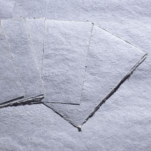 Puede incluir: Una pila de hojas de papel azul claro hechas a mano y texturizadas. El papel tiene una superficie rugosa e irregular y un borde rasgado. Las hojas est&aacute;n dispuestas en forma de abanico, con algunas superpuestas.