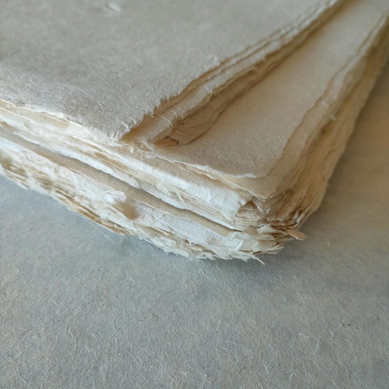 Op de afbeelding: Een stapel handgemaakt papier met ruwe, ongelijke randen. Het papier heeft een natuurlijke, gebroken witte kleur en een licht getextureerd oppervlak. De vellen zijn netjes opgestapeld, wat de unieke textuur en kwaliteit van het papier laat zien.