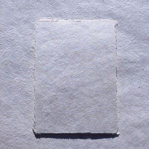 May include: A rectangular piece of handmade paper with a textured surface, in a light gray color. The paper has a deckled edge, and is set against a similar textured background.