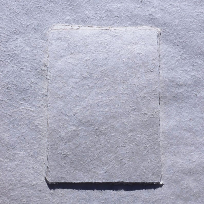 May include: A rectangular piece of handmade paper with a textured surface, in a light gray color. The paper has a deckled edge, and is set against a similar textured background.