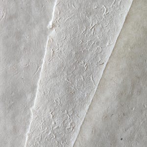 Op de afbeelding: Twee stukken getextureerd papier, een bovenop de ander. Het bovenste stuk papier heeft een ruw, onregelmatig oppervlak, terwijl het onderste stuk gladder is. Beide stukken papier zijn lichtbeige.