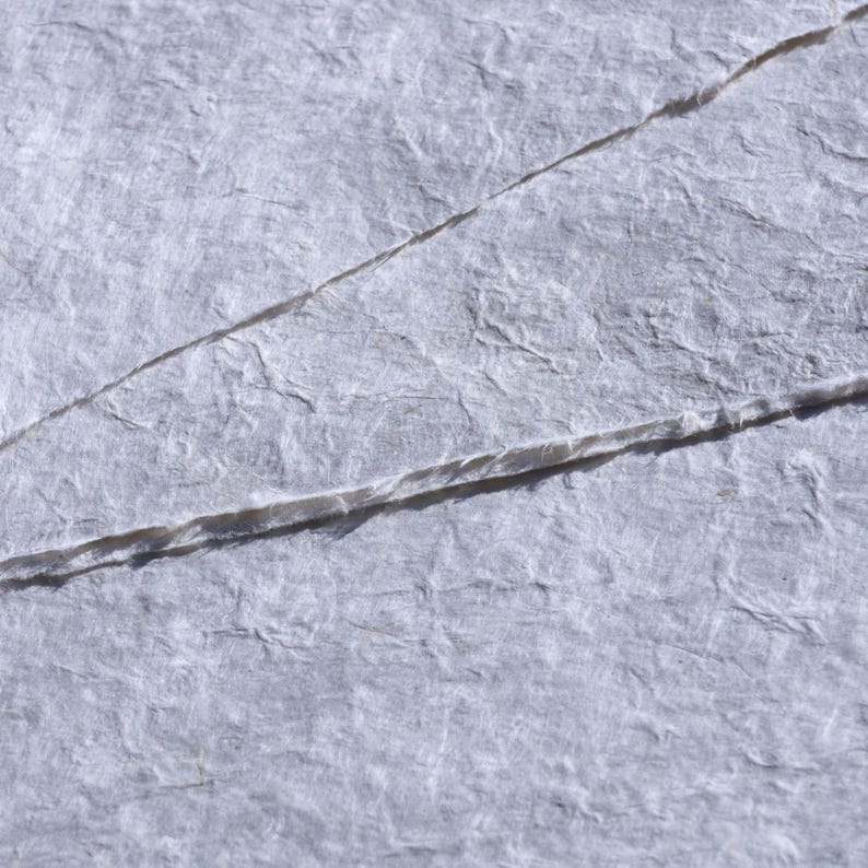 Pu&ograve; includere: Primo piano di carta strutturata grigio chiaro con fibre visibili e pieghe diagonali. La carta ha un aspetto ruvido e fatto a mano, suggerendo che potrebbe essere utilizzata per l'arte o l'artigianato.