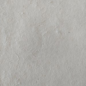 Op de afbeelding: Close-up van handgemaakt, gebroken wit papier met een getextureerd oppervlak. Het papier heeft zichtbare vezels en subtiele kleurvariaties, wat wijst op een natuurlijke, organische kwaliteit. Het oppervlak van het papier is licht ongelijkmatig.