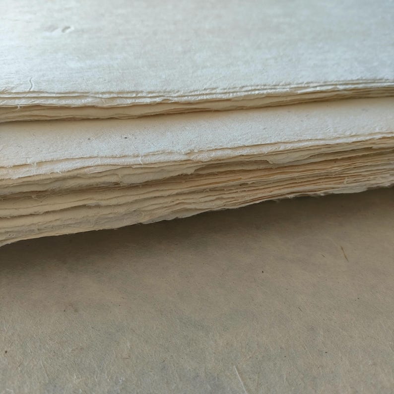 Op de afbeelding: Een stapel handgemaakte, cr&egrave;mekleurige papieren vellen met ruwe, gescheurde randen. Het papier heeft een licht getextureerd oppervlak en lijkt dik. De vellen zijn netjes opgestapeld, wat suggereert dat ze bedoeld zijn voor kunst- of schrijfprojecten.