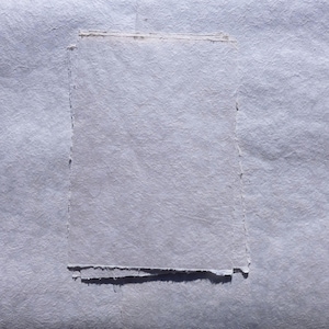 Puede incluir: Primer plano de papel hecho a mano de color blanco roto claro, con una superficie texturizada y fibras visibles. Los bordes son irregulares, lo que indica una fabricaci&oacute;n artesanal. El papel parece estar en capas.