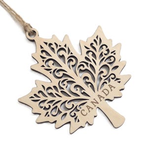 May include: A light-coloured wooden maple leaf ornament featuring intricate laser-cut scrollwork patterns throughout the surface. The word CANADA is engraved in capital letters along the lower right side. A thin jute twine loop is attached.