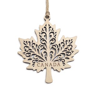 May include: A light-coloured wooden maple leaf ornament featuring intricate laser-cut scrollwork patterns throughout the leaf surface. The word CANADA is engraved in capital letters near the base of the stem. A thin jute twine loop is attached.