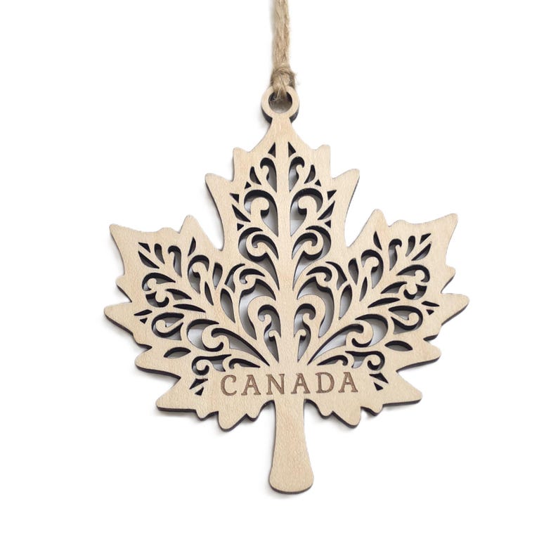 May include: A light-coloured wooden maple leaf ornament featuring intricate laser-cut scrollwork patterns throughout the leaf surface. The word CANADA is engraved in capital letters near the base of the stem. A thin jute twine loop is attached.