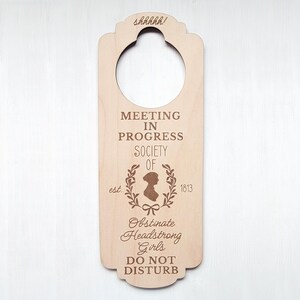 May include: Wooden door hanger with the text "Meeting in Progress Society of Obstinate Headstrong Girls est. 1813 Do Not Disturb" and a silhouette of a woman with a wreath around her head.