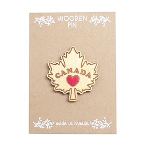 May include: Wooden maple leaf pin with "CANADA" and a red heart. The pin is on a brown card with "WOODEN PIN" and "made in canada". The pin is a symbol of Canada.