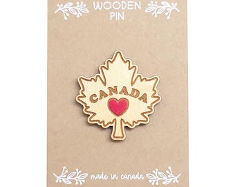 Puede incluir: Pin de madera con forma de hoja de arce con la palabra "CANADA" y un corazón rojo. La tarjeta marrón lleva las palabras "WOODEN PIN" y "made in canada".