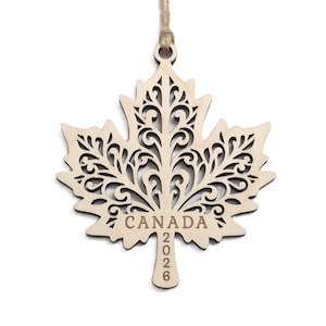 May include: A light-coloured wooden maple leaf ornament featuring intricate laser-cut scrollwork patterns. The word CANADA is engraved horizontally across the centre, with the year 2026 engraved vertically on the stem. A jute twine loop is attached.