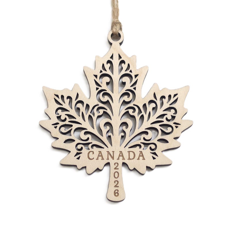 May include: A light-coloured wooden maple leaf ornament featuring intricate laser-cut scrollwork patterns. The word CANADA is engraved horizontally across the centre, with the year 2026 engraved vertically on the stem. A jute twine loop is attached.