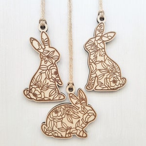 May include: Three wooden bunny ornaments with a floral design. Each bunny is attached to a twine string for hanging.