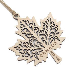 May include: A light-coloured wooden maple leaf ornament featuring intricate laser-cut scrollwork patterns throughout the leaf. The word CANADA and the year 2026 are engraved on the stem. A thin jute twine loop is attached to the top for hanging.