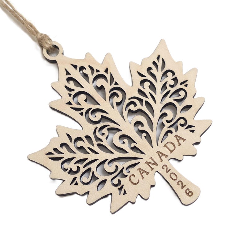 May include: A light-coloured wooden maple leaf ornament featuring intricate laser-cut scrollwork patterns throughout the leaf. The word CANADA and the year 2026 are engraved on the stem. A thin jute twine loop is attached to the top for hanging.