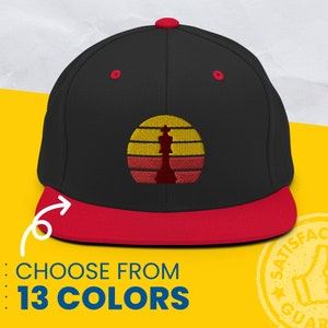 Puede incluir: Gorra de béisbol negra y roja con un gráfico de puesta de sol rojo y amarillo y una silueta de pieza de ajedrez. Elige entre 13 colores.