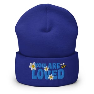 May include: A royal blue beanie with a white and yellow embroidered design that says "You Are Loved" with daisies and a bee.