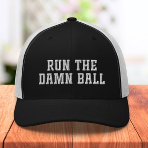 May include: Black and white trucker hat with a white mesh back. The hat has the text "RUN THE DAMN BALL" embroidered in white on the front.
