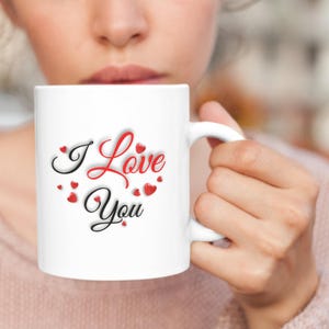 A woman is drinking coffee from a white Valentines mug that features the message I Love You which is written in red and black letters all surrounded by small hearts.  A wonderful Valentines Day, Christmas or Birthday gift for Mom or Dad, Boyfriend or Girlfriend.