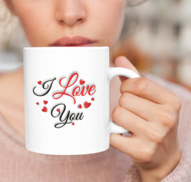 A woman is drinking coffee from a white Valentines mug that features the message I Love You which is written in red and black letters all surrounded by small hearts.  A wonderful Valentines Day, Christmas or Birthday gift for Mom or Dad, Boyfriend or Girlfriend.