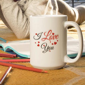 A child is lying on the floor coloring, beside her is a white ceramic mug with the words I Love You, the lettering is written in red and black letters all surrounded by many small hearts. I love you gift for him or her